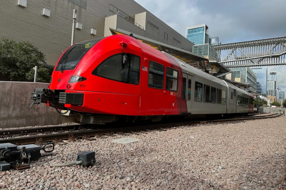Red modern train on tracks with large warehouse-type building behind it in Austin, Texas.